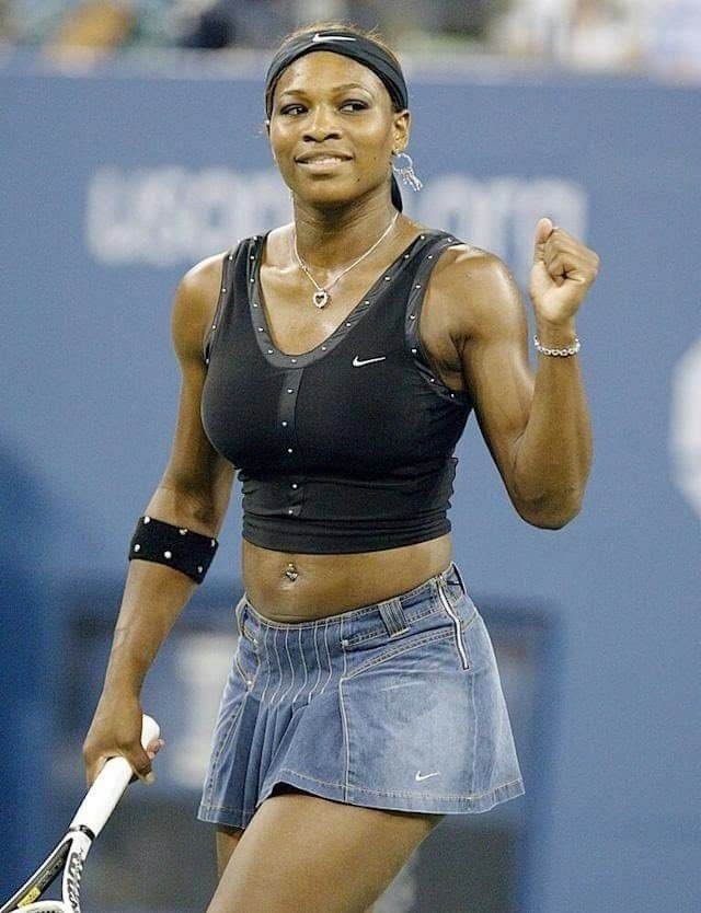Serena Williams tennis player wearing a black athletic crop top and a denim tennis skirt, holding a racquet and raising her fist in celebration on a hard-court stadium wearing a Tennis Bracelet.