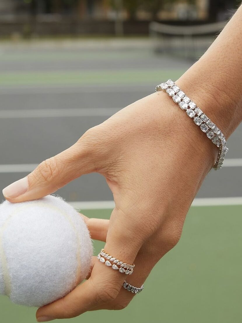 A close-up of a hand holding a tennis ball on a court, adorned with a diamond tennis bracelet and stacked diamond rings, highlighting the sporty elegance and origin of tennis jewelry styling.