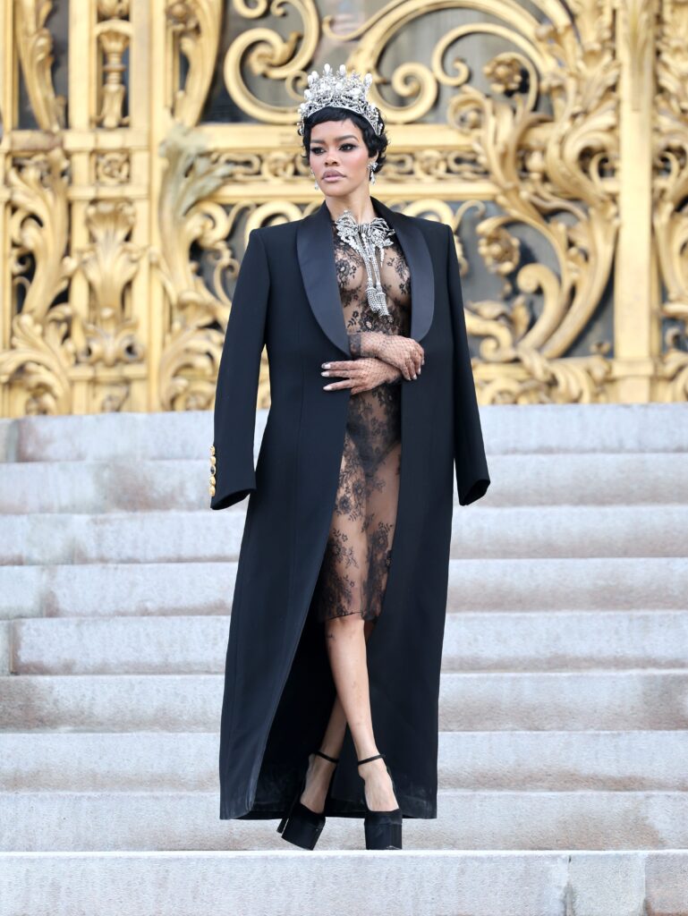 Teyana Taylor wearing a pearl and diamond style tiara and dramatic bow necklace at Paris Haute Couture Week, standing on the steps of the Petit Palais in front of ornate gold gates, dressed in a sheer black lace outfit and long black coat, inspired by historic French crown jewels.
