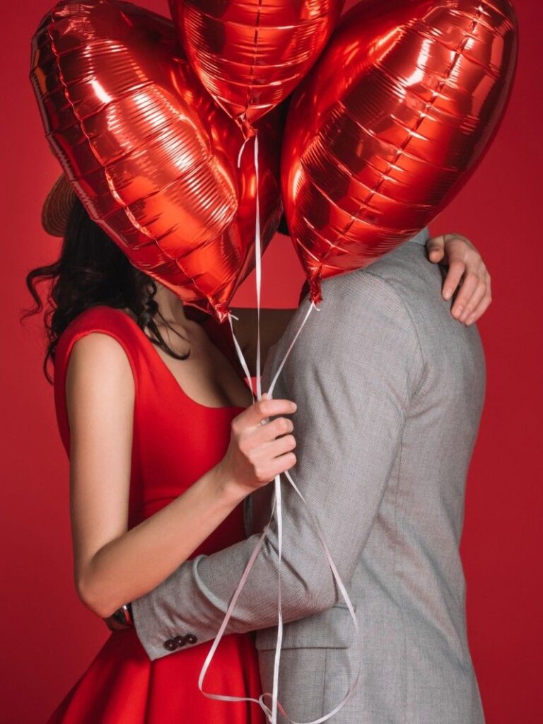 Romantic couple embracing against a red backdrop, faces hidden behind heart-shaped balloons, symbolising modern Valentine’s Day love and emotional connection.