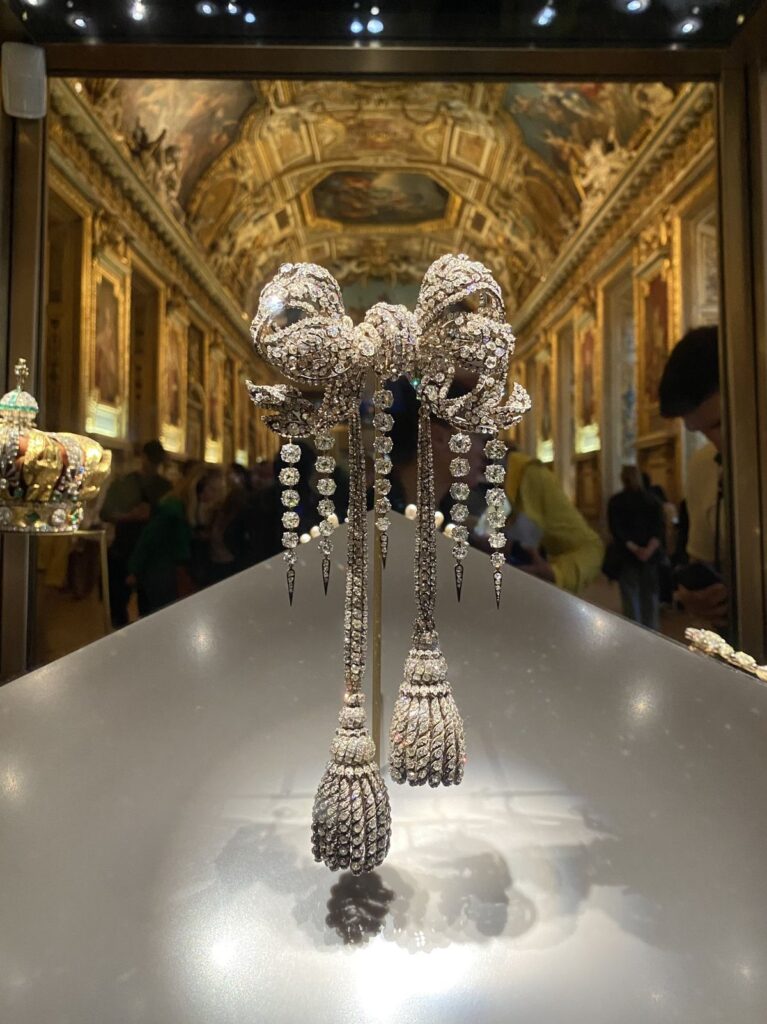 Historic diamond bow brooch with long tassel drops displayed inside a museum gallery, crafted in 19th-century French royal style and associated with crown jewel collections.