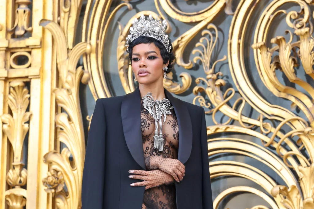 Teyana Taylor wearing a pearl and diamond style tiara and dramatic bow necklace at Paris Haute Couture Week, standing on the steps of the Petit Palais in front of ornate gold gates, dressed in a sheer black lace outfit and long black coat, inspired by historic French crown jewels.
