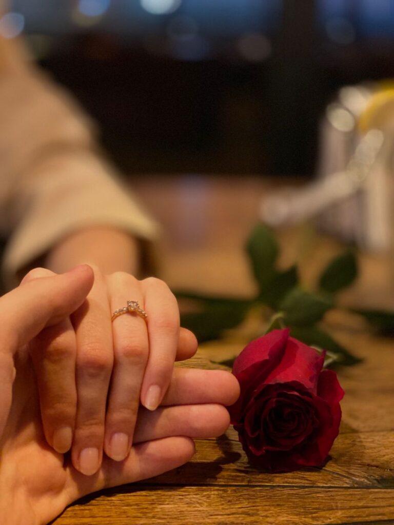 Close-up of hands holding an engagement ring beside a red rose on a wooden table, symbolising timeless love, commitment, and Valentine’s Day traditions.
